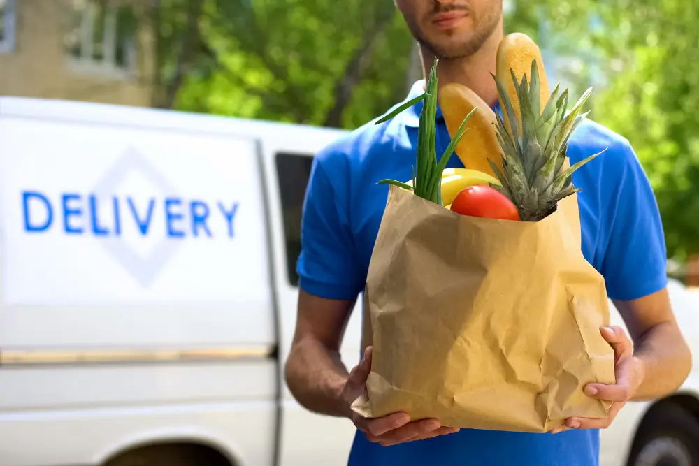 A courier in blue holding a paper bag of groceries in front of a van lettered Delivery