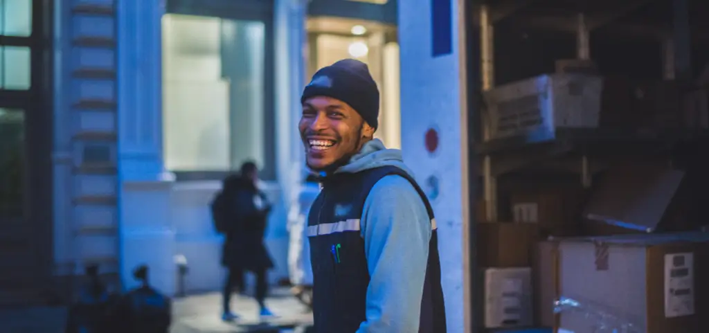 A smiling delivery worker in a toque and reflective vest standing beside an open truck loaded with boxes on a city street at dusk