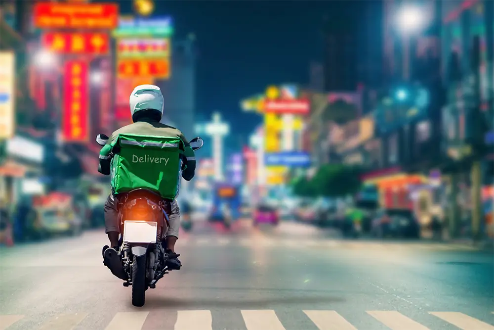 A motorcycle courier with a green Delivery box on the back riding down a city street lit by neon signs at night