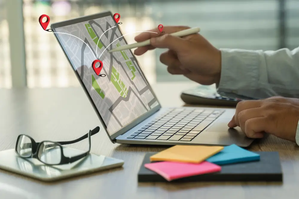 A laptop showing a street map with map pins rising from the screen, linked by route lines