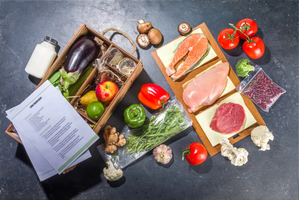 A meal kit laid out on a dark counter: a crate of produce, salmon, chicken and beef on a board, loose vegetables and printed recipe cards