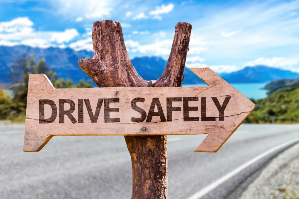 A wooden arrow sign reading Drive Safely beside a road, mountains behind