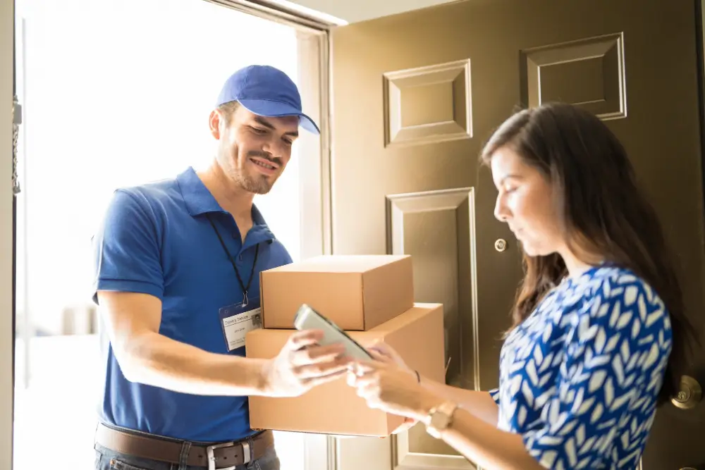 A courier holding two boxes at a front door while the recipient signs for them on a phone