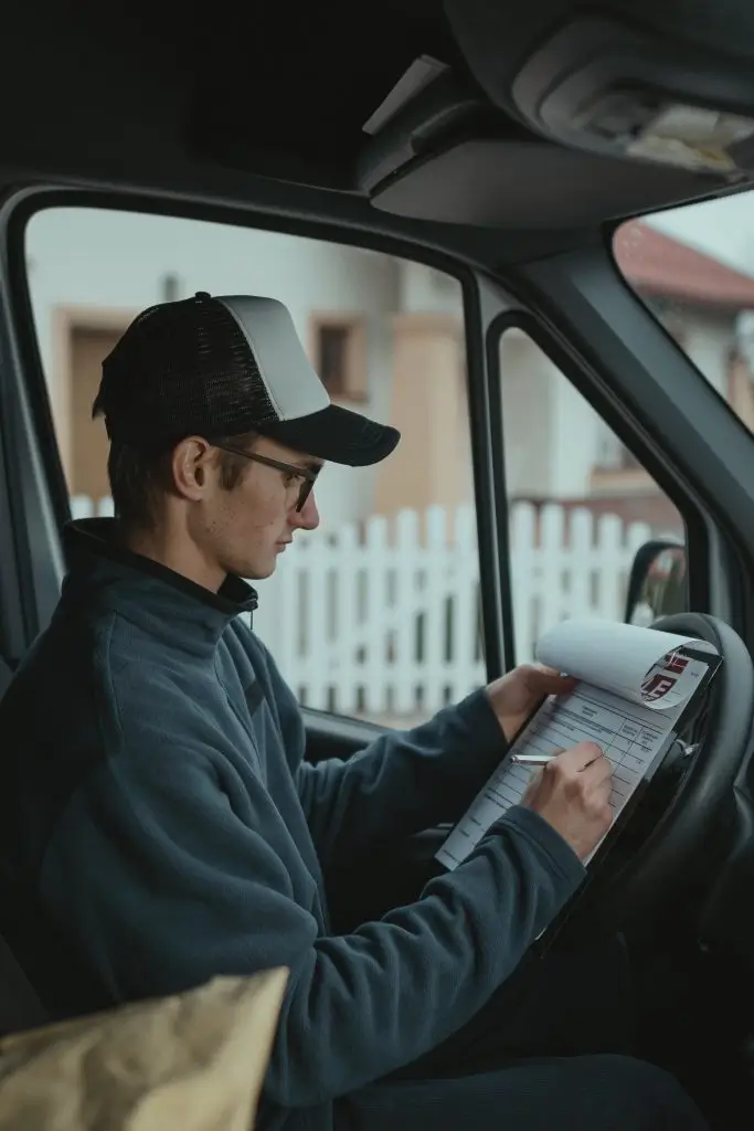 A driver in a cap sitting in a van’s driver’s seat, filling in a paper log on a clipboard propped on the steering wheel