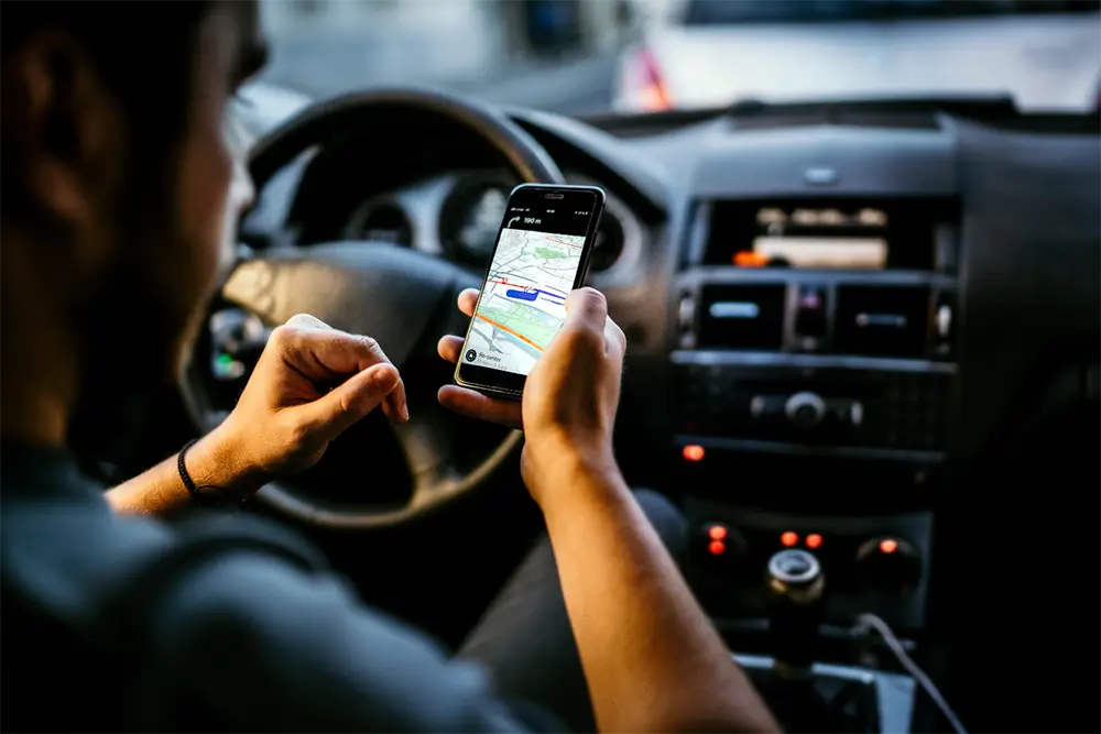 A driver behind the wheel of a car holding a phone showing a navigation map with the route highlighted