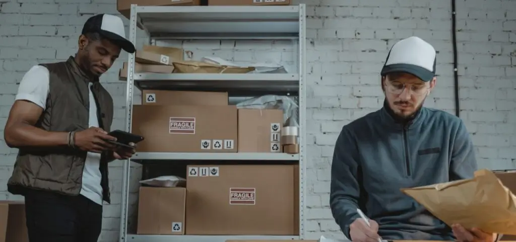 Two workers in a stockroom, one scanning with a handheld device and one labelling an envelope, shelves of cartons between them