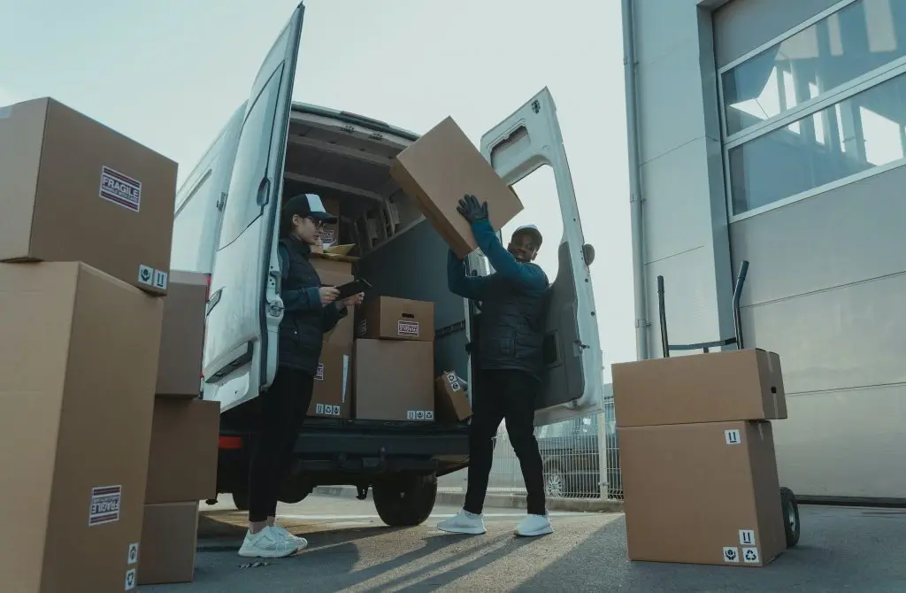 Two workers unloading cartons from a van outside a depot, one checking a handheld device