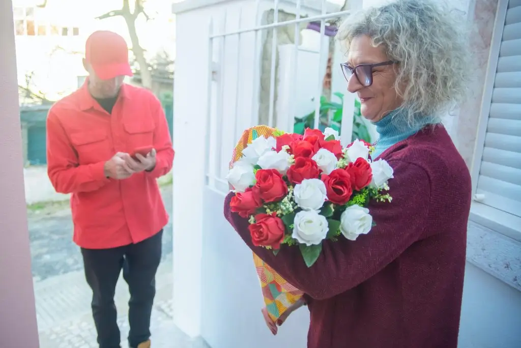 A woman at her door holding a wrapped bouquet of red and white roses while the courier checks his phone