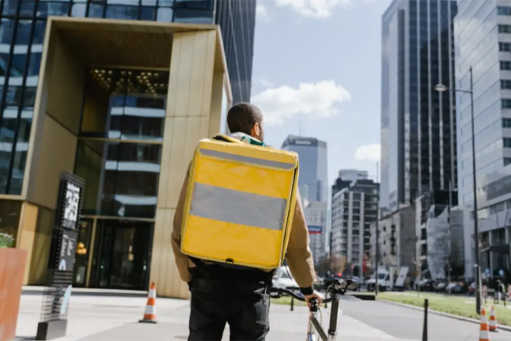 A cycle courier seen from behind with a large yellow insulated backpack, walking a bike through a city street