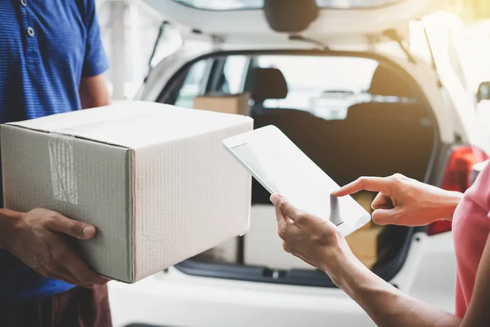 A courier holding out a carton while a customer signs on a tablet at the back of a car